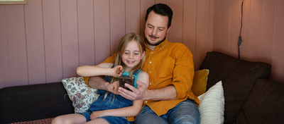 Foto av far og datter som ser på en mobil og smiler. De sitter i en sofa, og hun er i armkroken hans. 