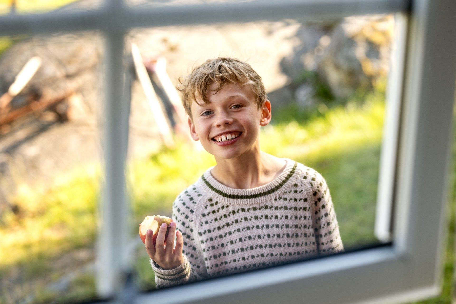 Gutt ser inn gjennom vindu, smiler og spiser eple. Foto
