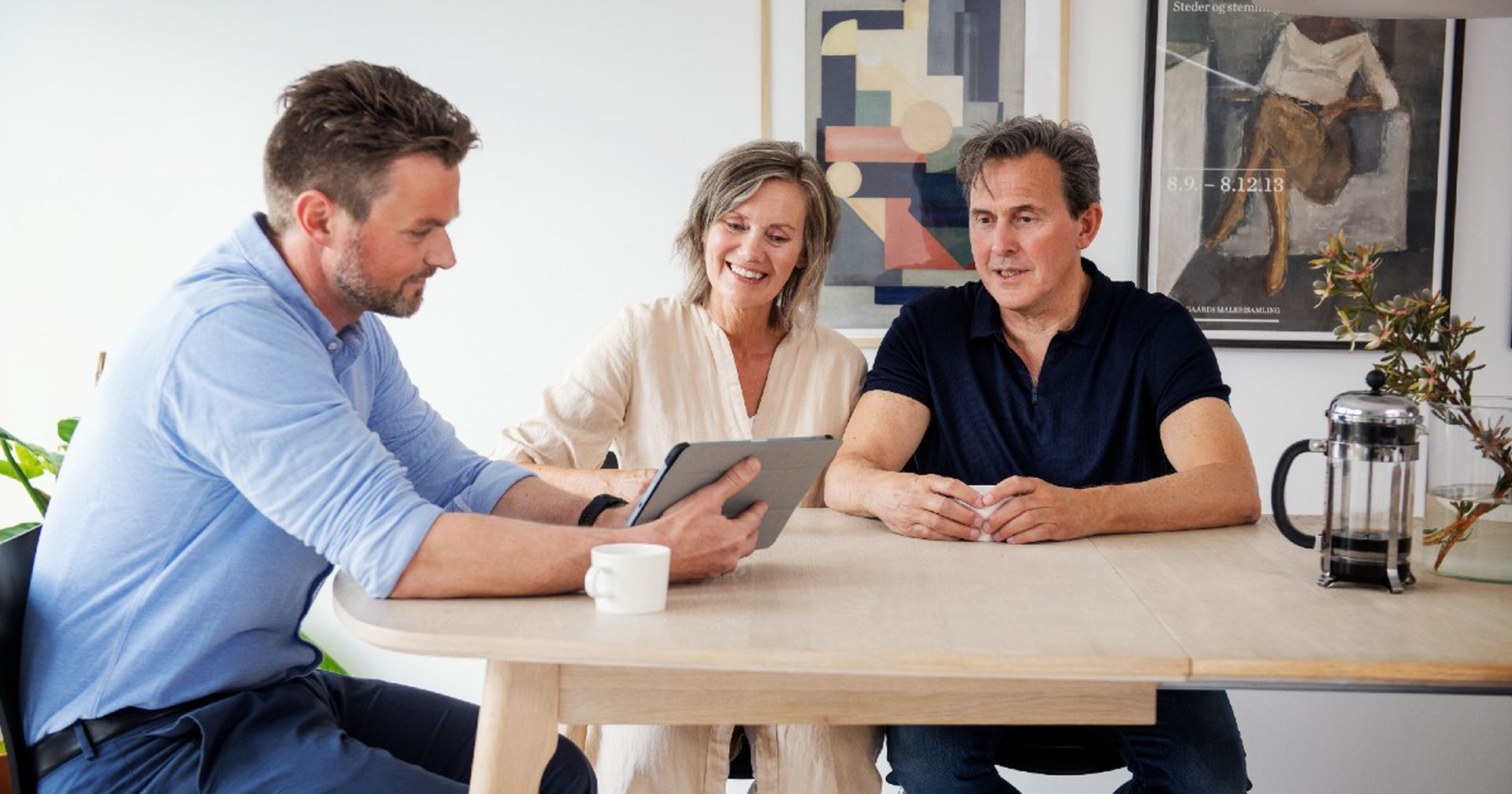 Par sitter sammen ved et bord i møte med en eiendomsmegler som viser frem noe på en tablet. Foto.