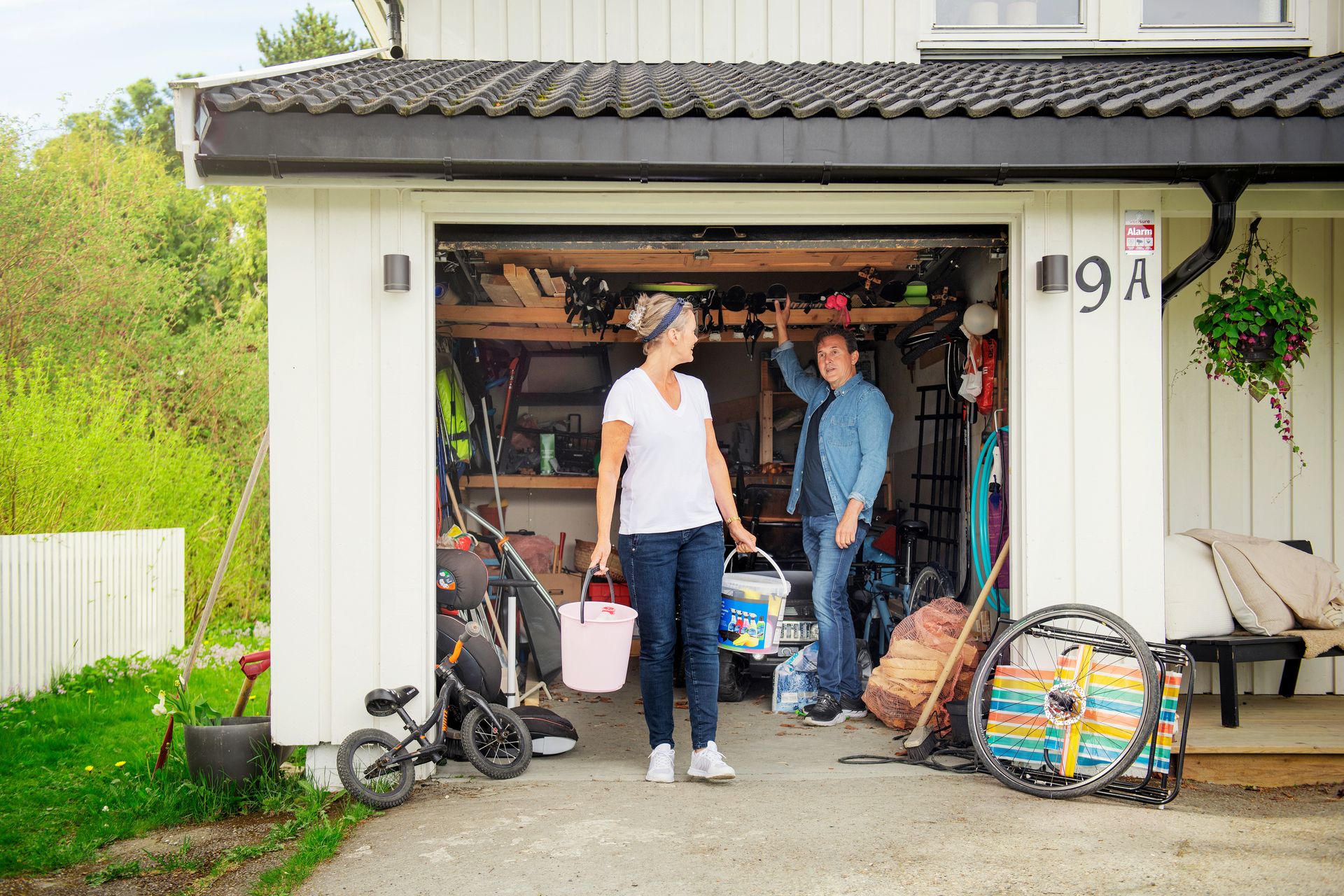 Par står i åpen garasjeport og snakker, kvinne bærer to bøtter. Foto.
