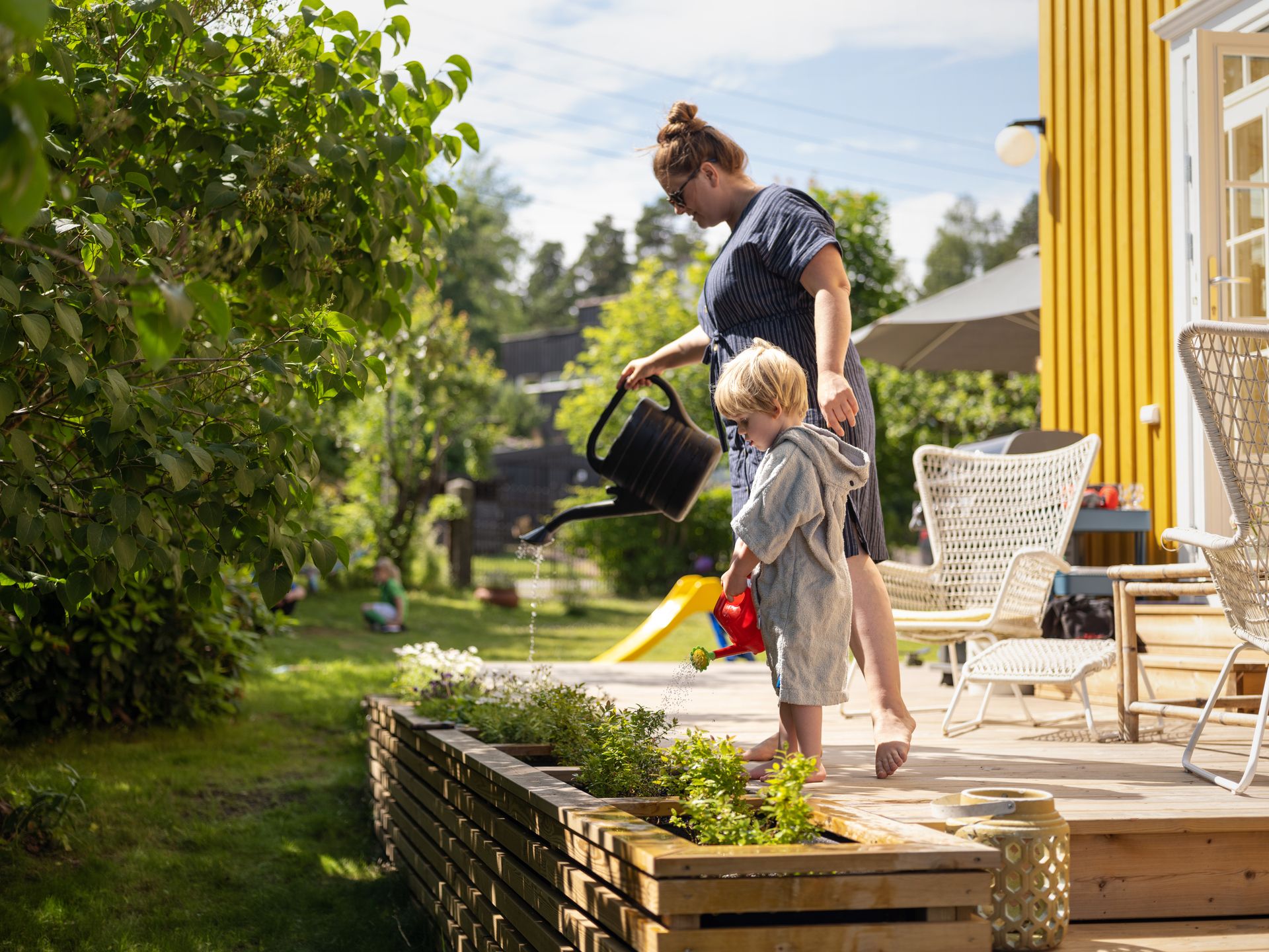Hagevanning.jpg Foto av en dame og et barn foran et gult hus. De står på en terrasse og vanner grønne planter fra hver sin kanne.