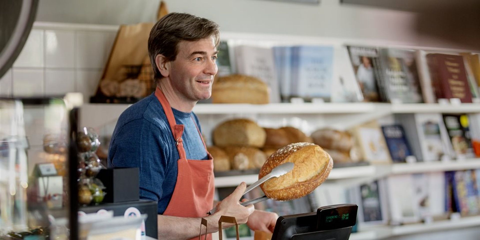 Mann bak kasse på bakeri holder et brød med klype og smiler. Bilde