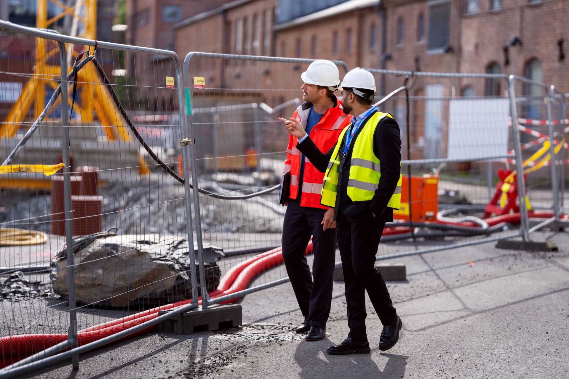 To personer med hjelm og refleksvest går på en byggeplass, ved et gjerde. Foto
