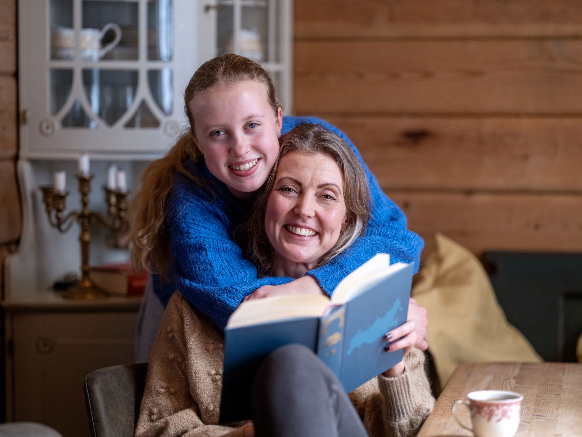 Mor sitter ved et bord og leser bok, datter gir klem bakfra. Begge smiler mot kamera. Foto
