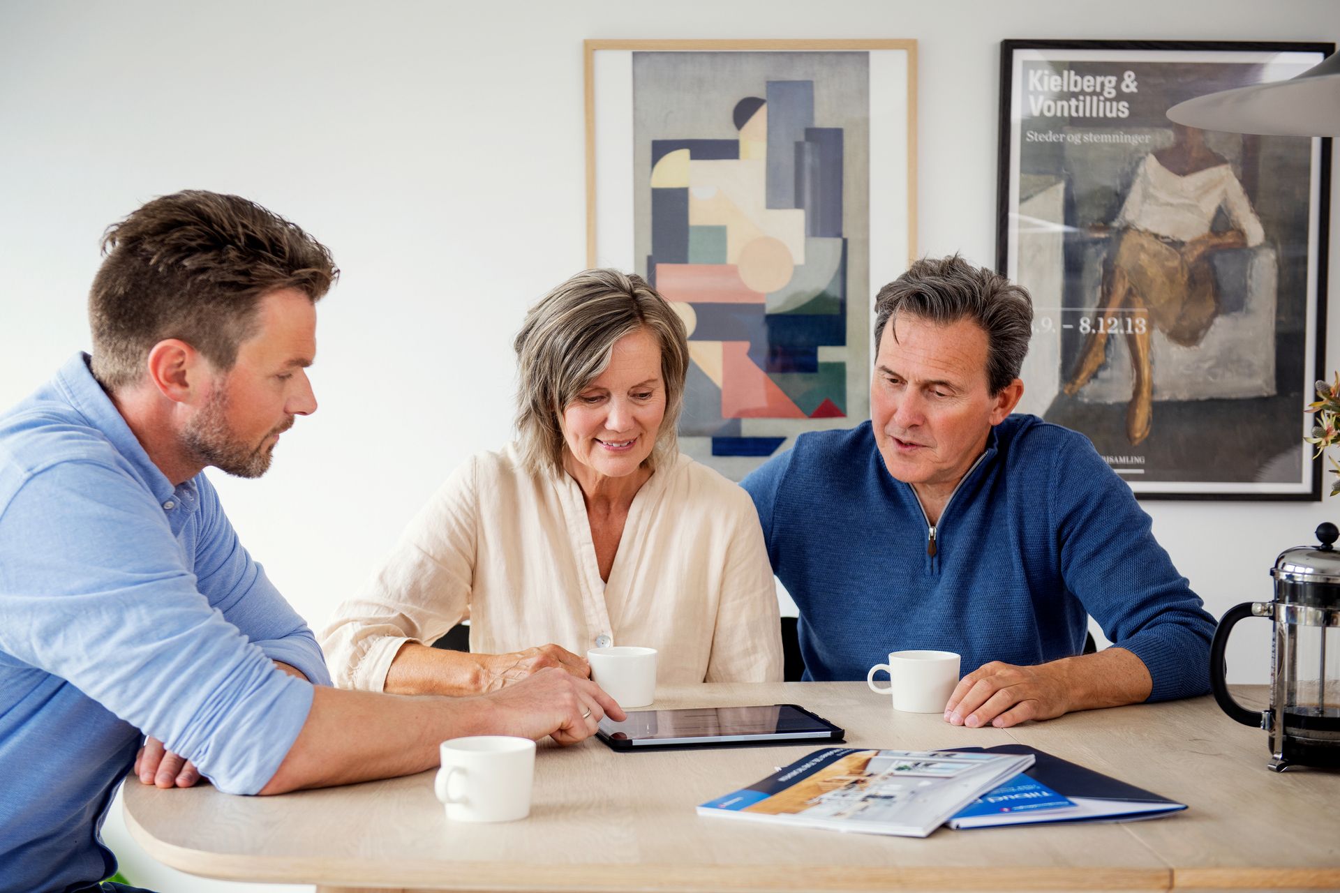 Par sitter sammen ved et bord i møte med en eiendomsmegler som peker på noe på en tablet. Foto.
