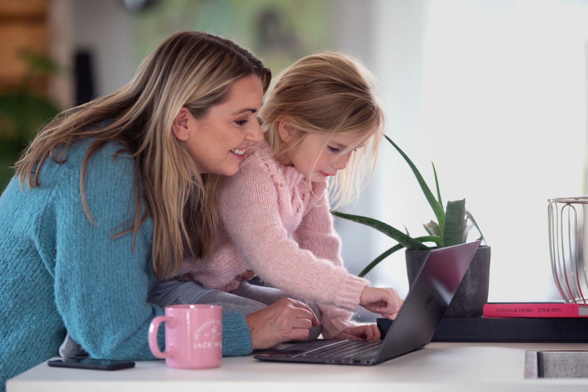 Foto av kvinne og barn. Kvinnen står ved en kjøkkenbenk, mens jenta sitter på den. Begge ser på en PC-skjerm. 