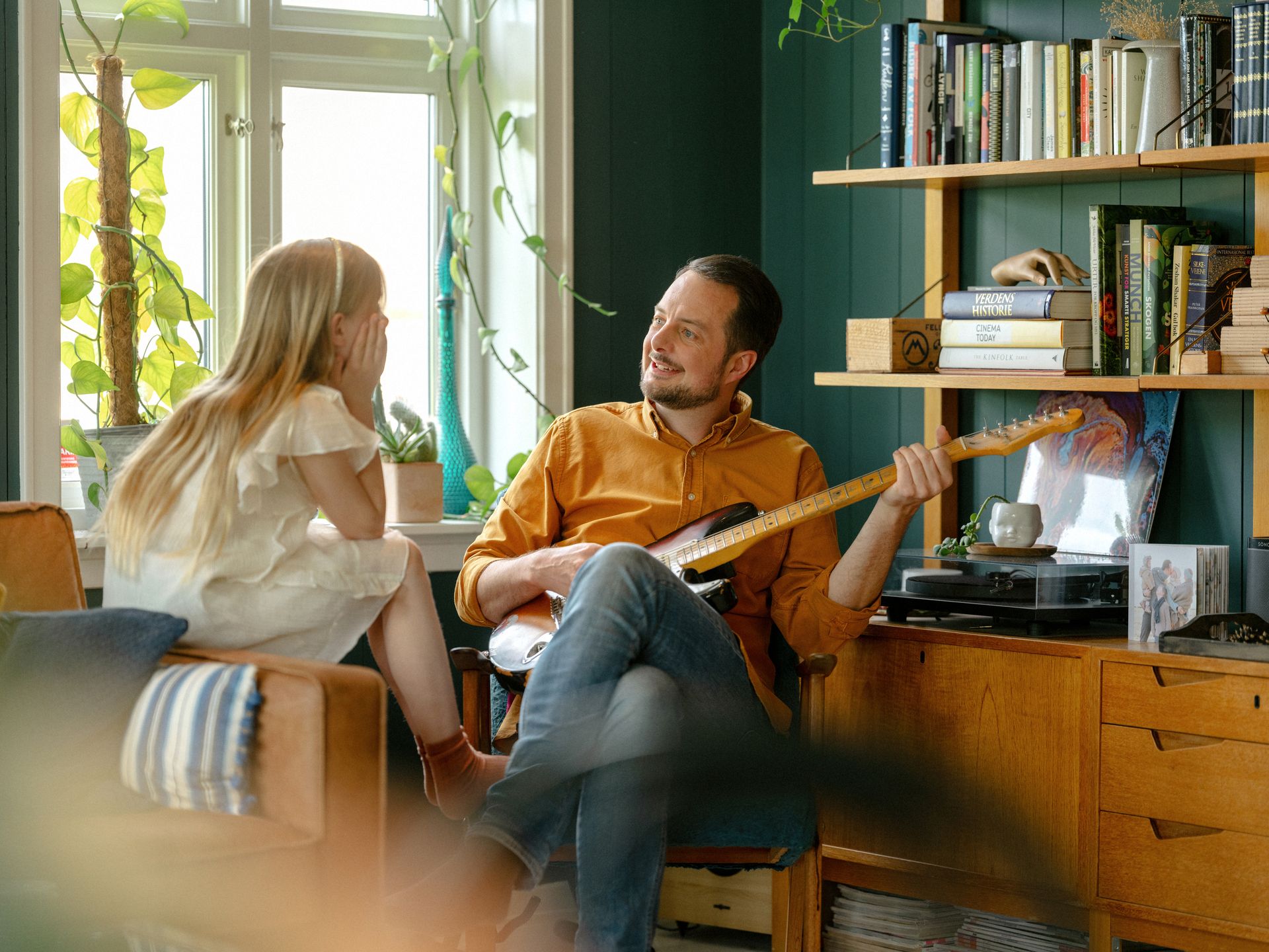 Foto av en pappa som spiller gitar for datteren sin. Han sitter i en stol, og hun på kanten av en sofa. De ser på hverandre