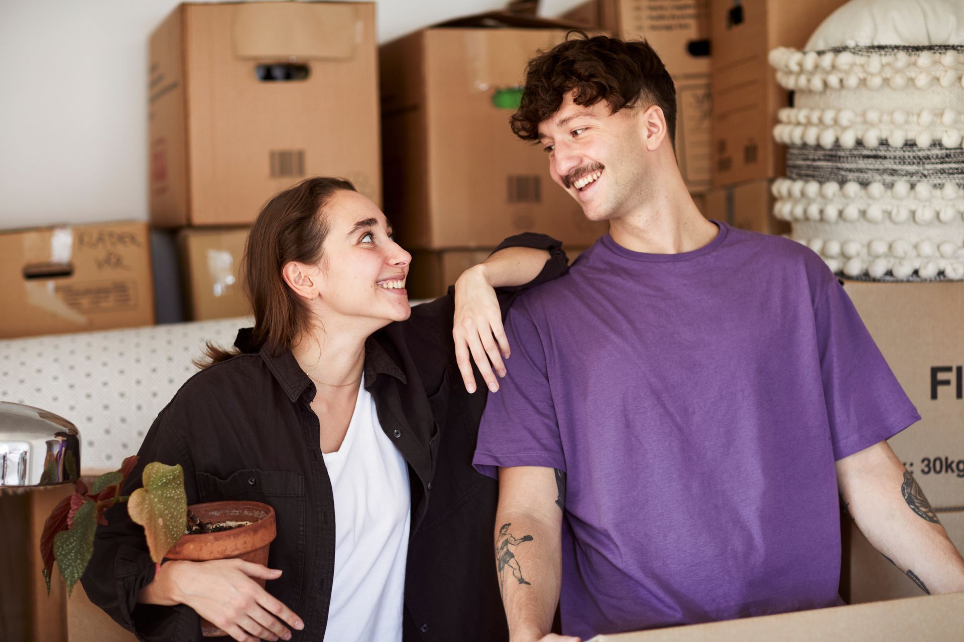 Foto av mann og kvinne som er på flyttefot. De smiler til hverandre, og i bakgrunnen er det stablet mange pappesker