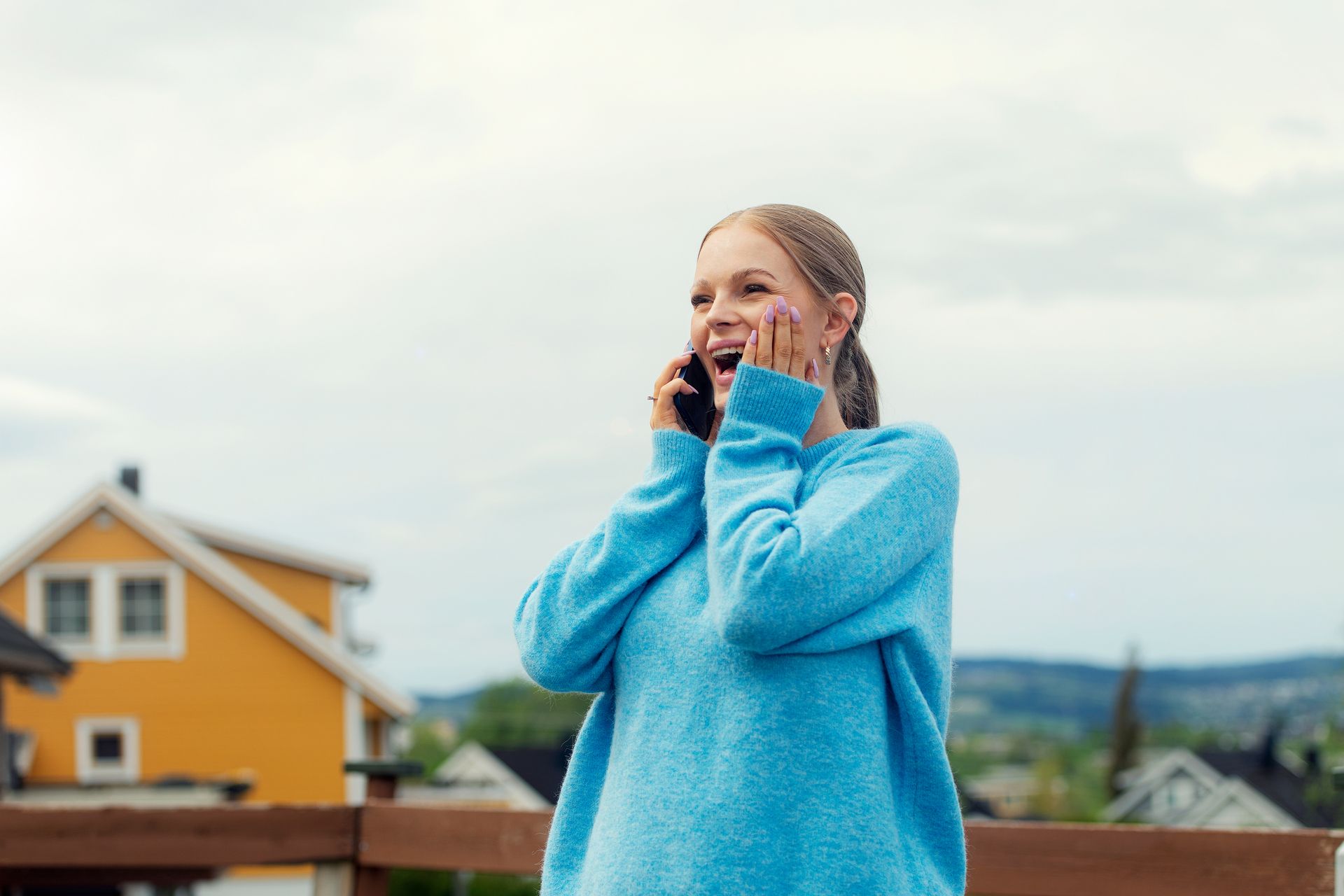 Kvinne snakker i telefon, smiler bredt og tar seg til ansiktet. Foto.

