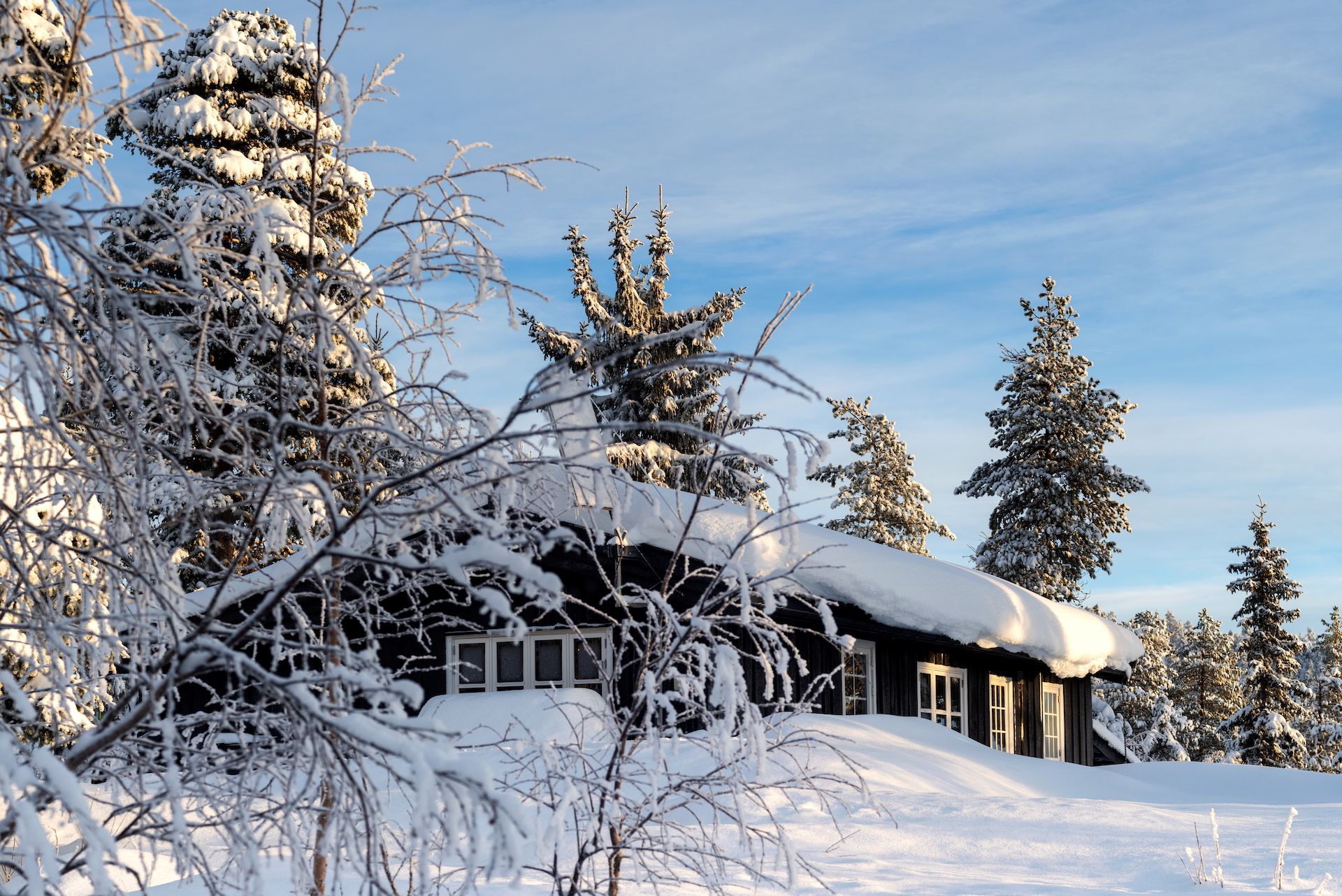 Hytte i skogen med snø på taket og på tunet. Det er blå himmel, og solen skinner på hytta. Foto. 