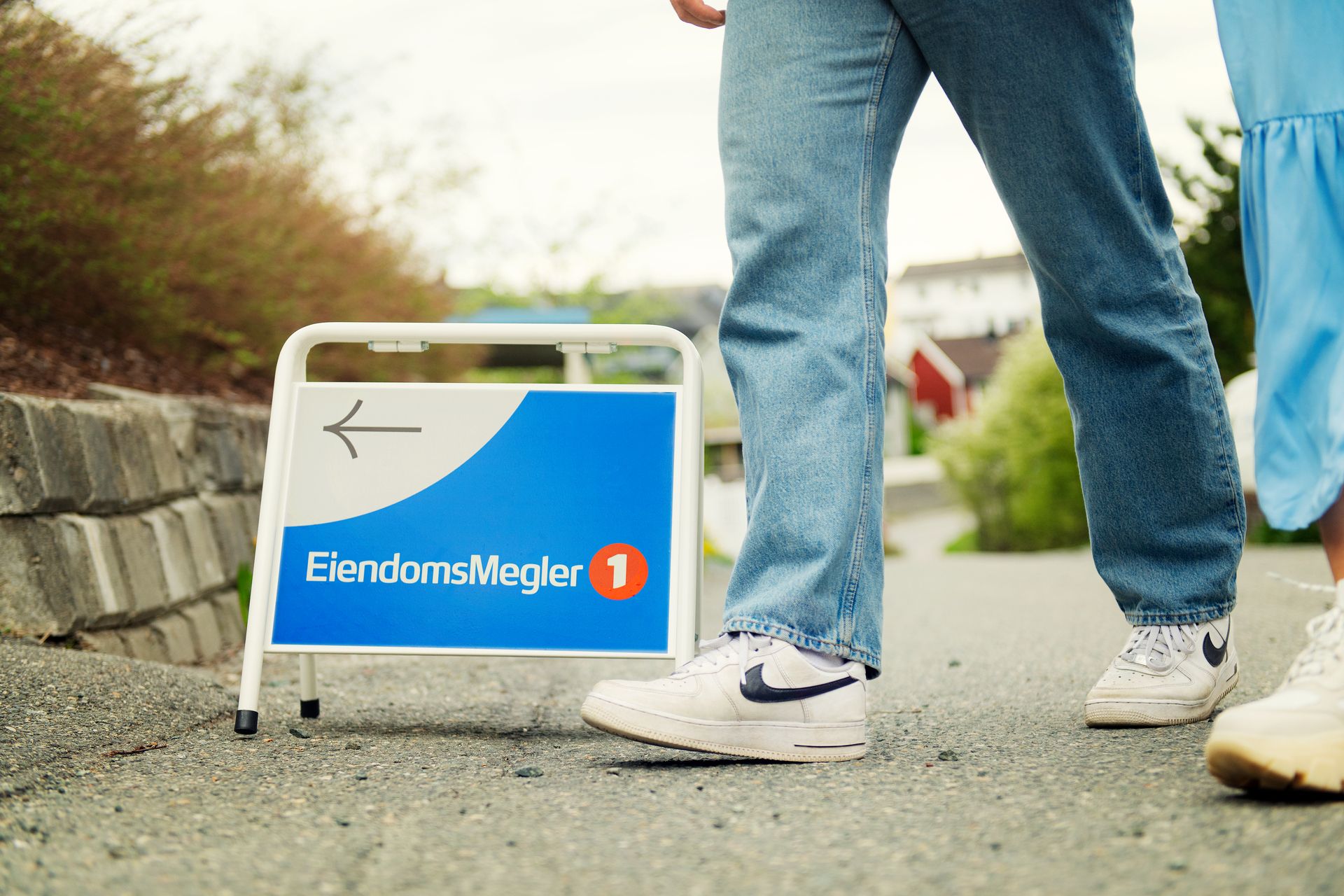Beina til to personer passerer et visningsskilt med EiendomsMegler 1-logo. Foto.
