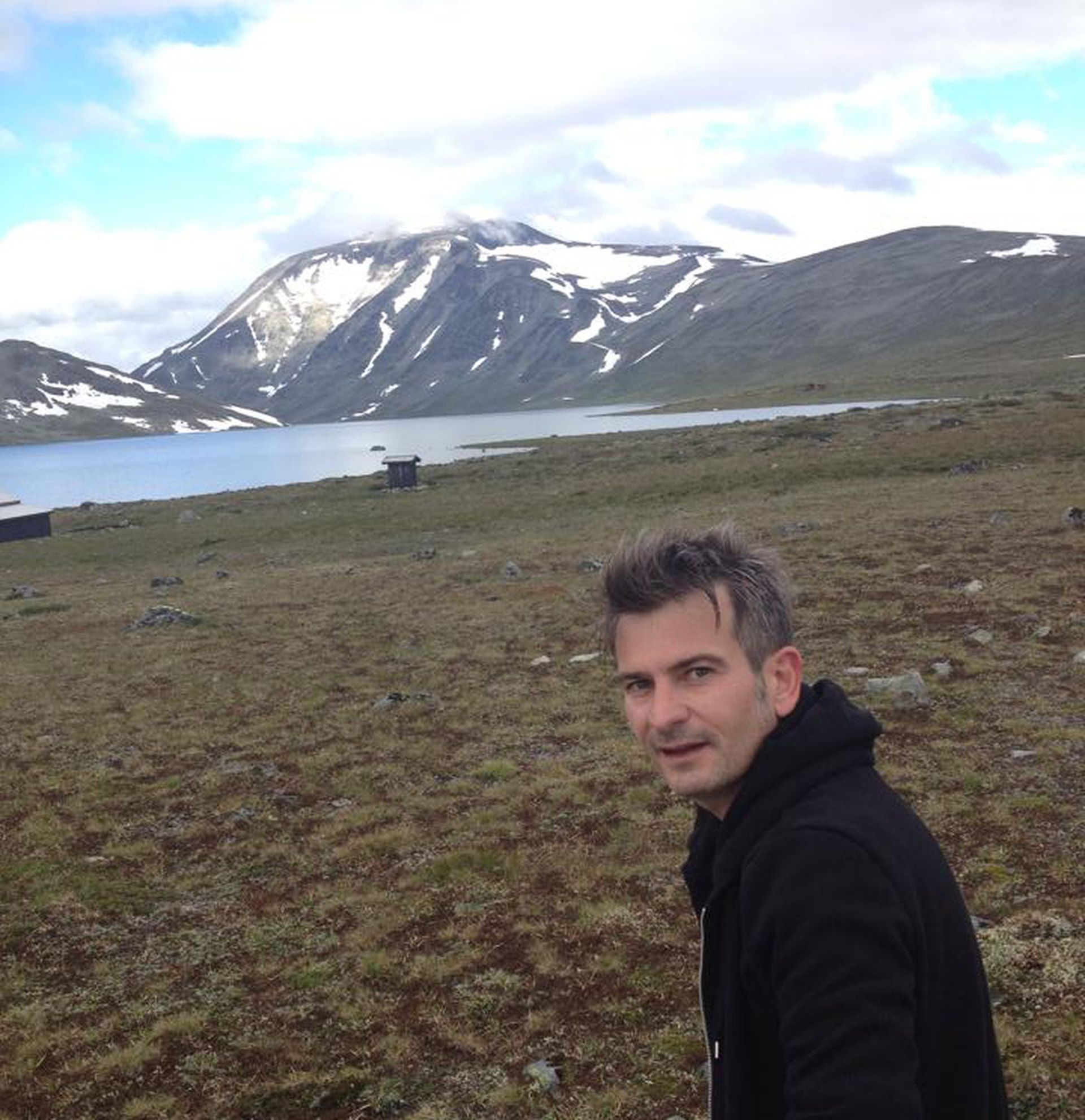 Mann i fjell-landskap med innsjø og store snøkledde fjell i bakgrunnen. Bilde.