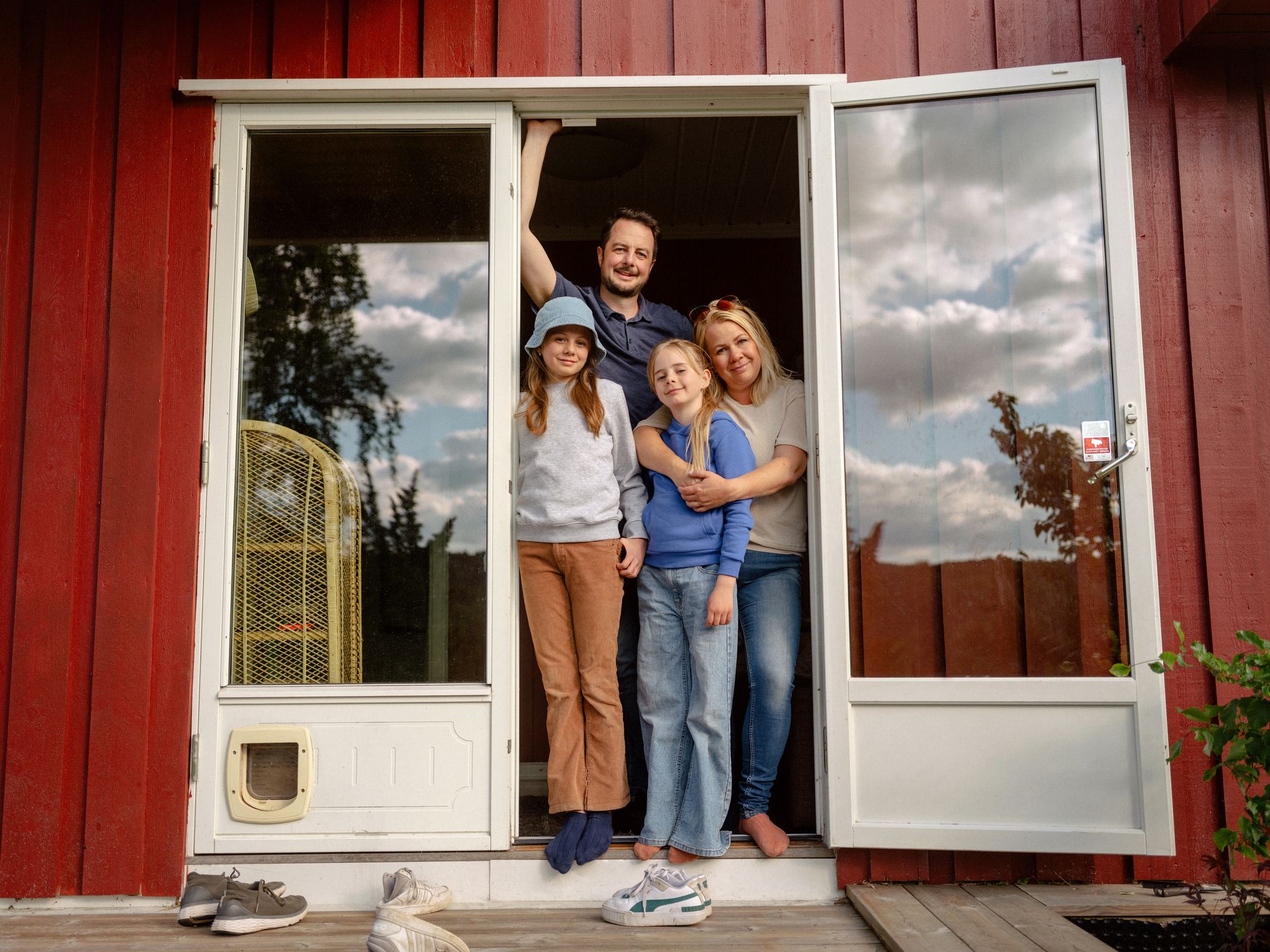 Foto av en familie med far, mor og do døtre som står i en åpen terrassedør og smiler til kamera.