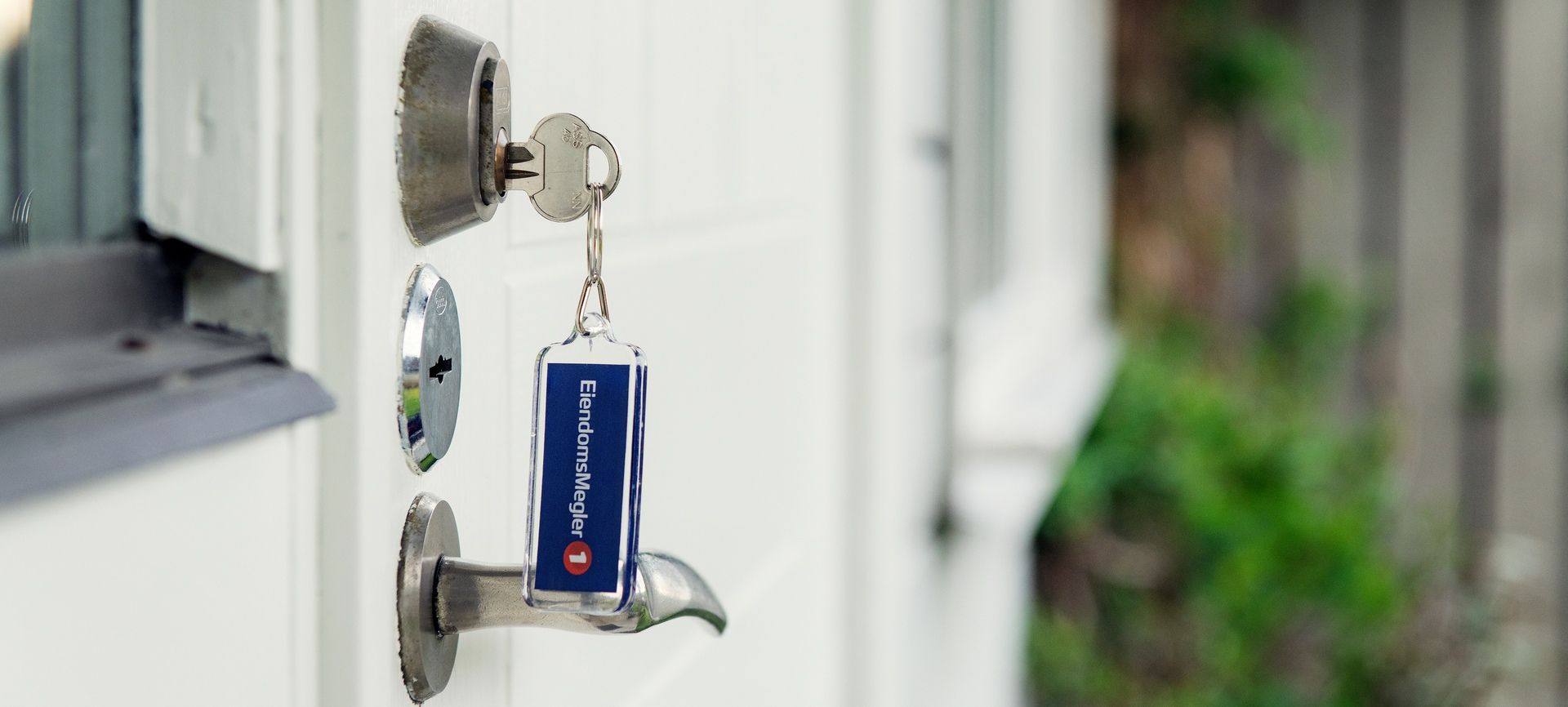 Nøkkel står i låsen på en hvit dør. Blå nøkkelring med EiendomsMegler 1-logo. Foto