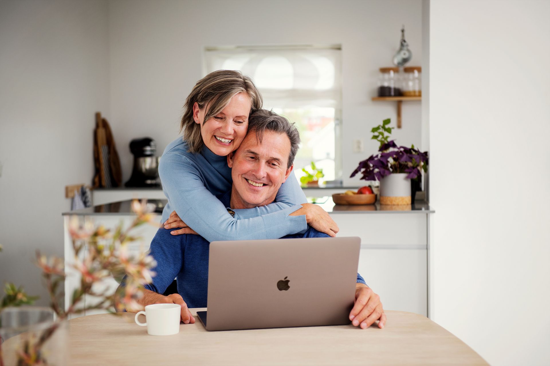 Voksent par smiler og ser på en mac-skjerm, dame holder rundt mann. Foto.

