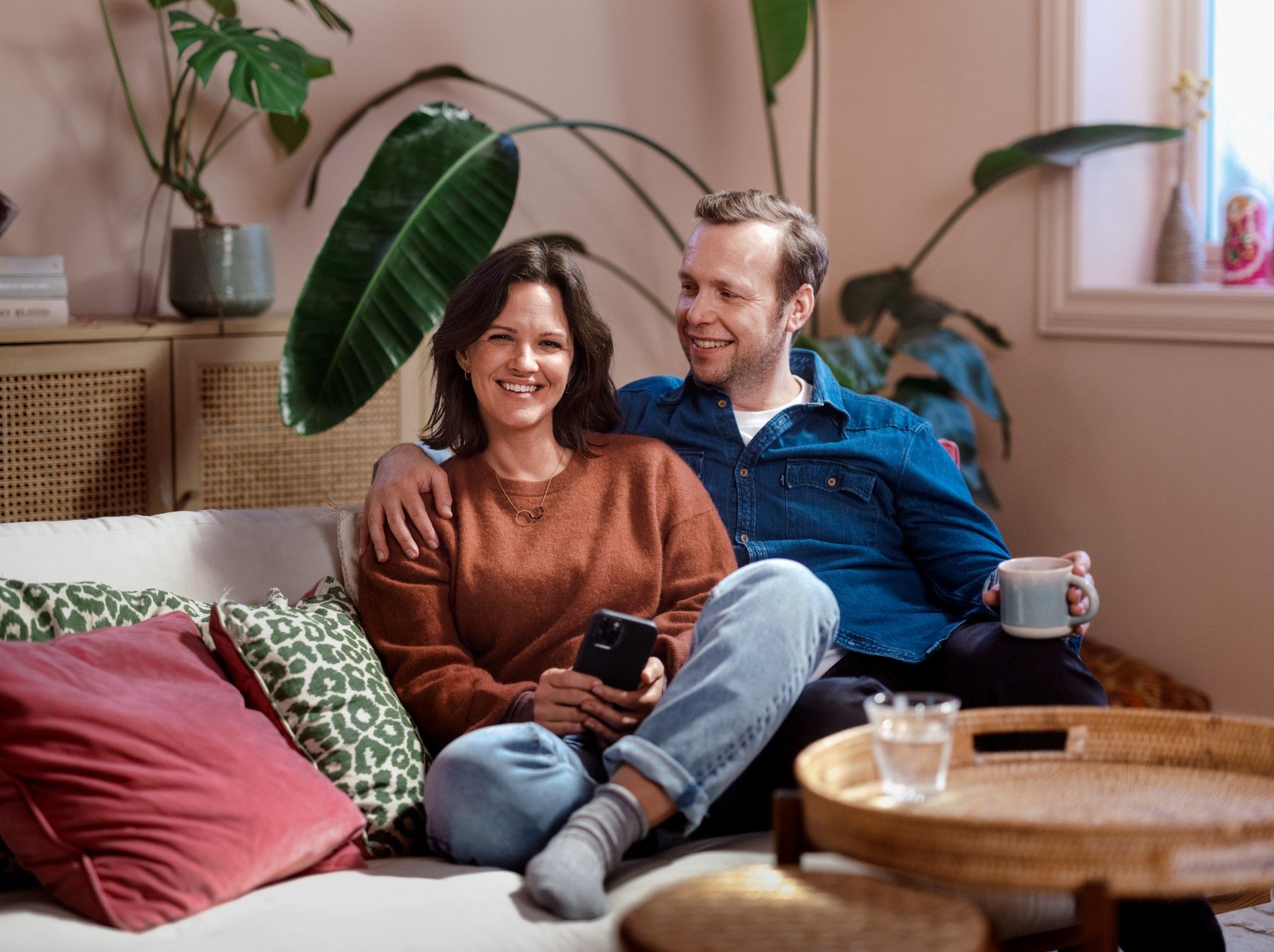 Kvinne og mann sitter i sofa, vendt mot kamera. Møbler og planter i bakgrunnen. Foto
