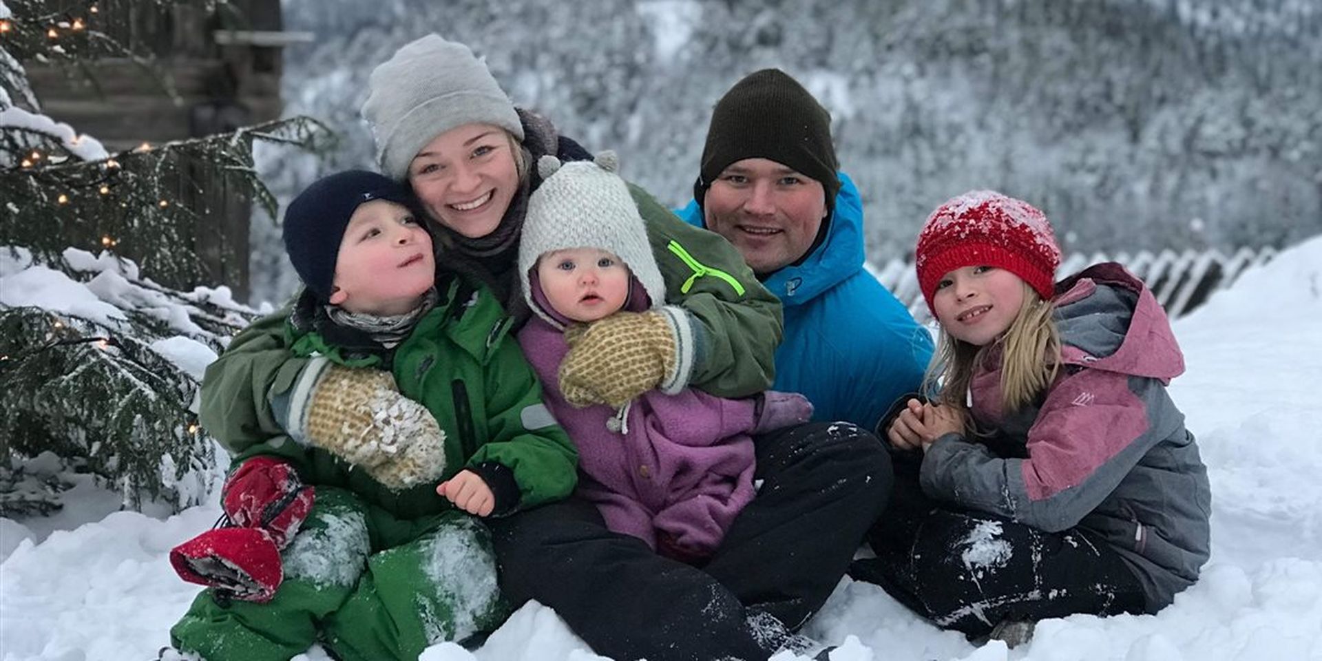 To voksne og tre barn sitter i snøen i vinterklær, ser på kamera. Bilde