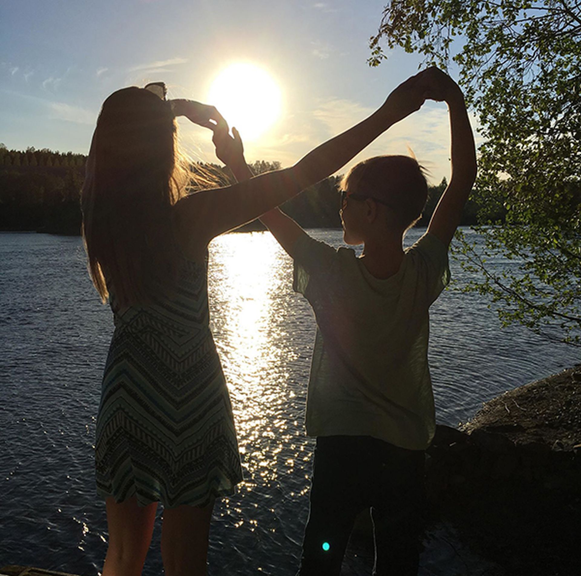 To barn holder hender. Står med ryggen til kamera, ser mot solnedgang ved vann. Bilde.