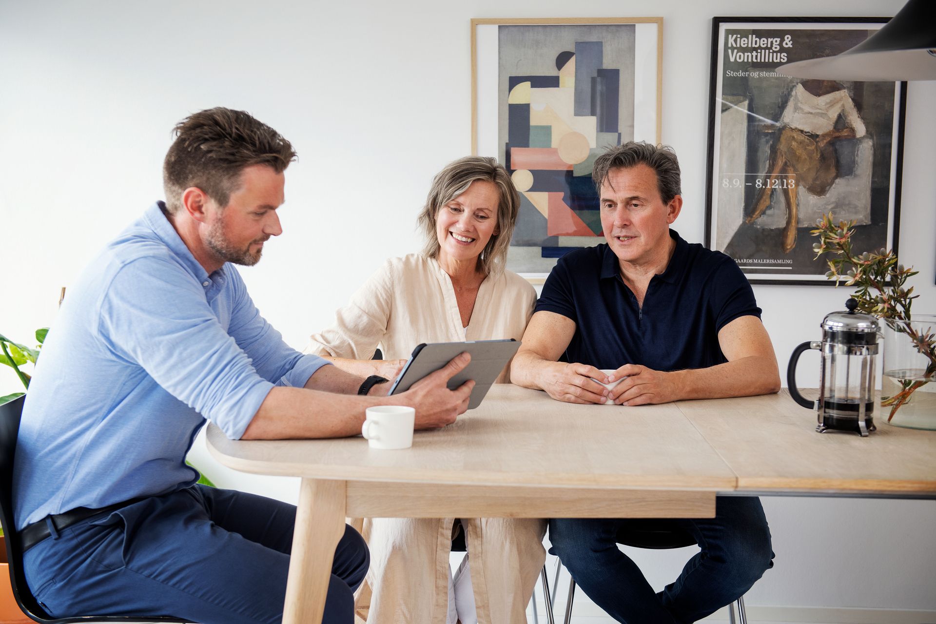 Par sitter sammen ved et bord i møte med en eiendomsmegler som peker på noe på en tablet. Foto.
