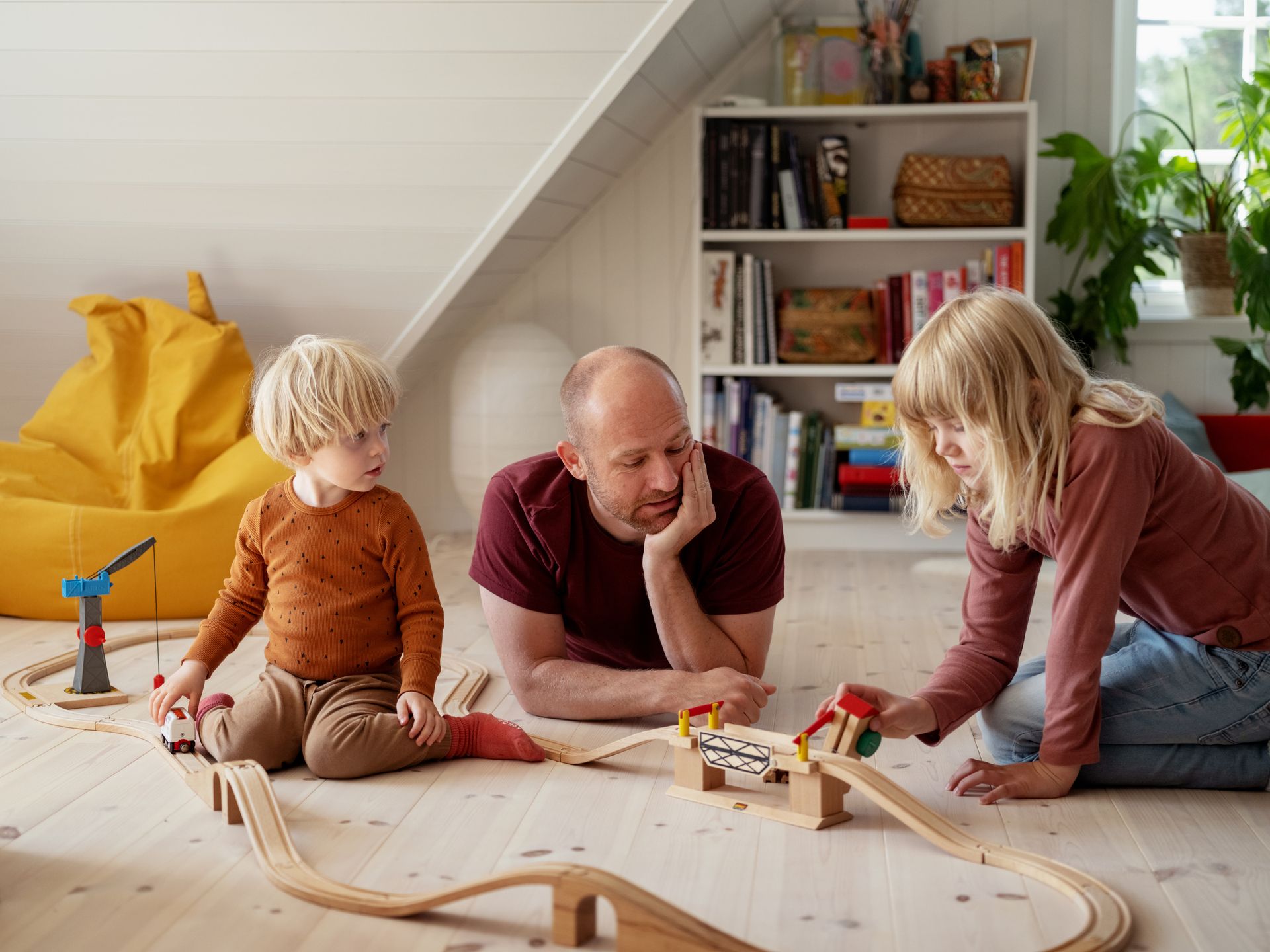 Foto av lillebror, storesøster og pappa som leker med tog på barnerommet. 
