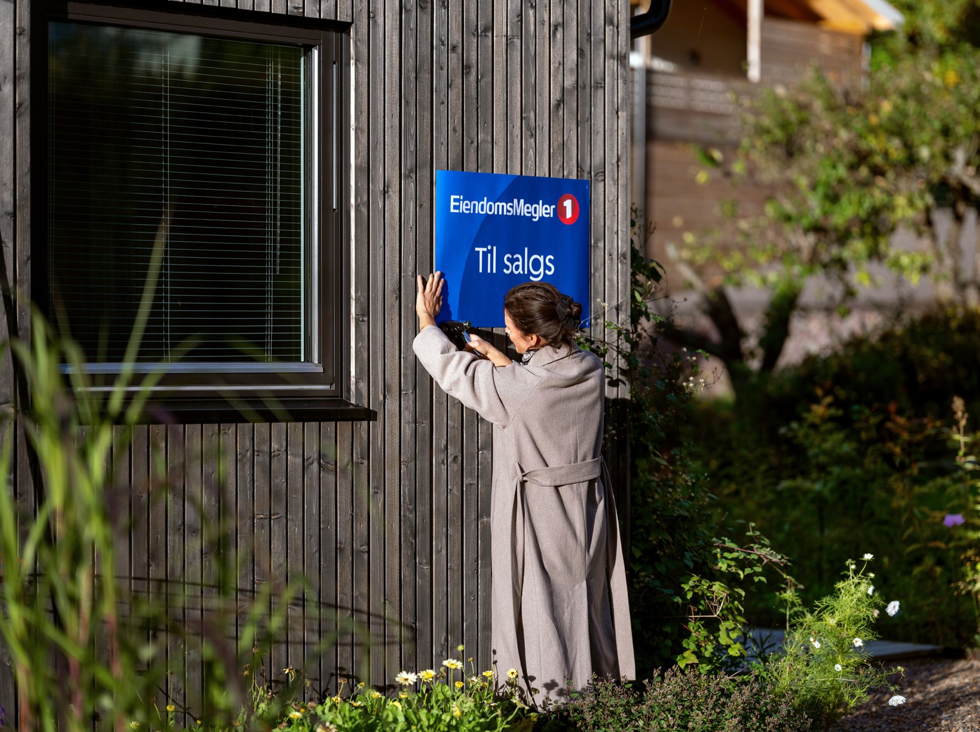 Dame henger opp en til salgs-plakat på en trefarget husvegg. Foto
