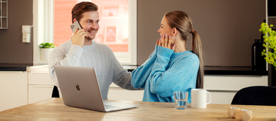 Foto av mann og kvinne som sitter ved et spisebord med en Mac oppå. Han smiler og snakker i telefonen, og kvinnen ser glad ut.