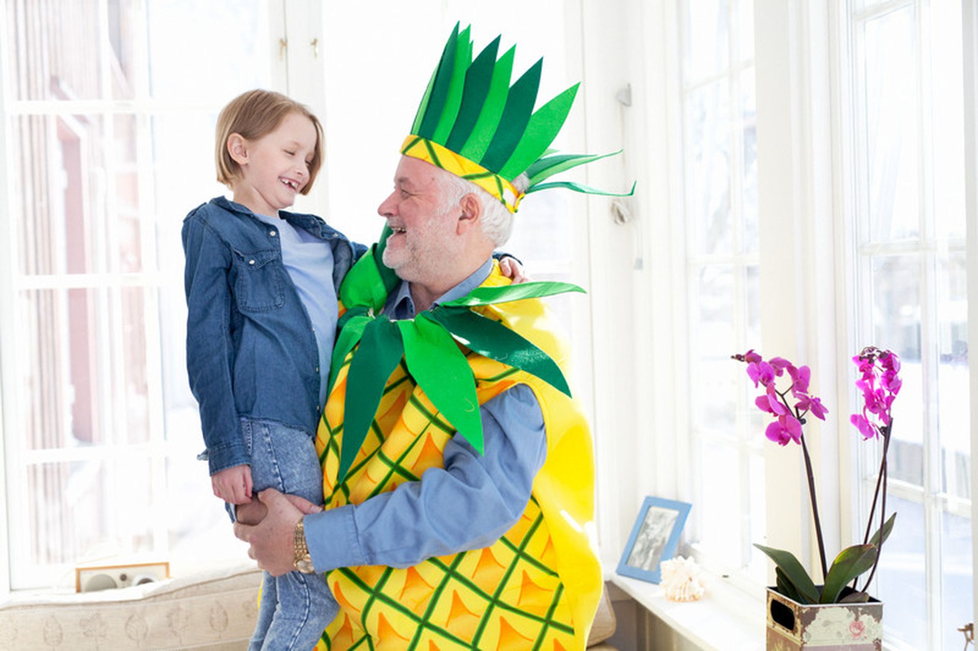 Bestefar i ananaskostyme løfter opp et barn. Bilde.