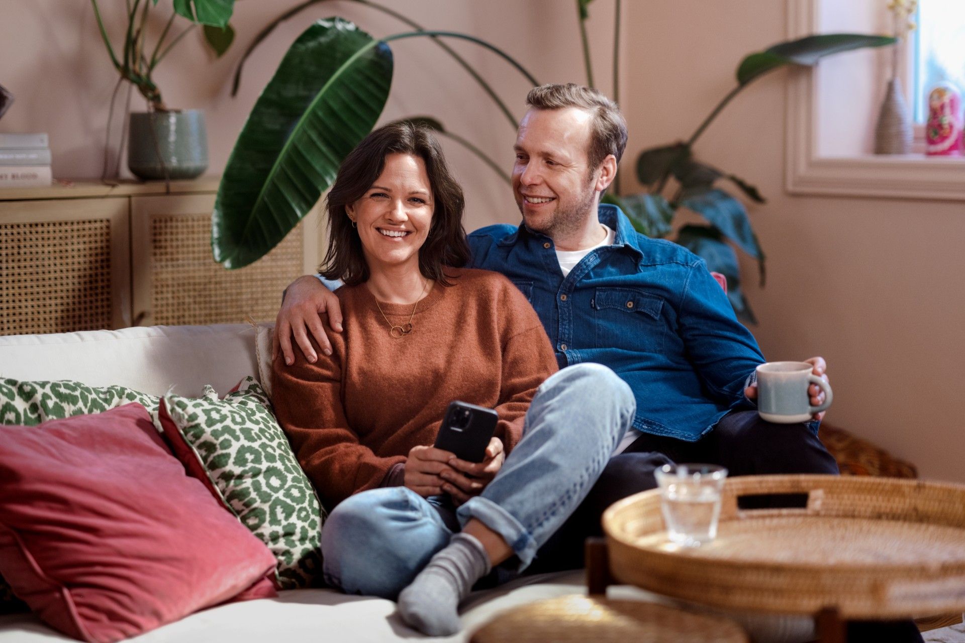 Kvinne og mann sitter i sofa, vendt mot kamera. Møbler og planter i bakgrunnen. Foto
