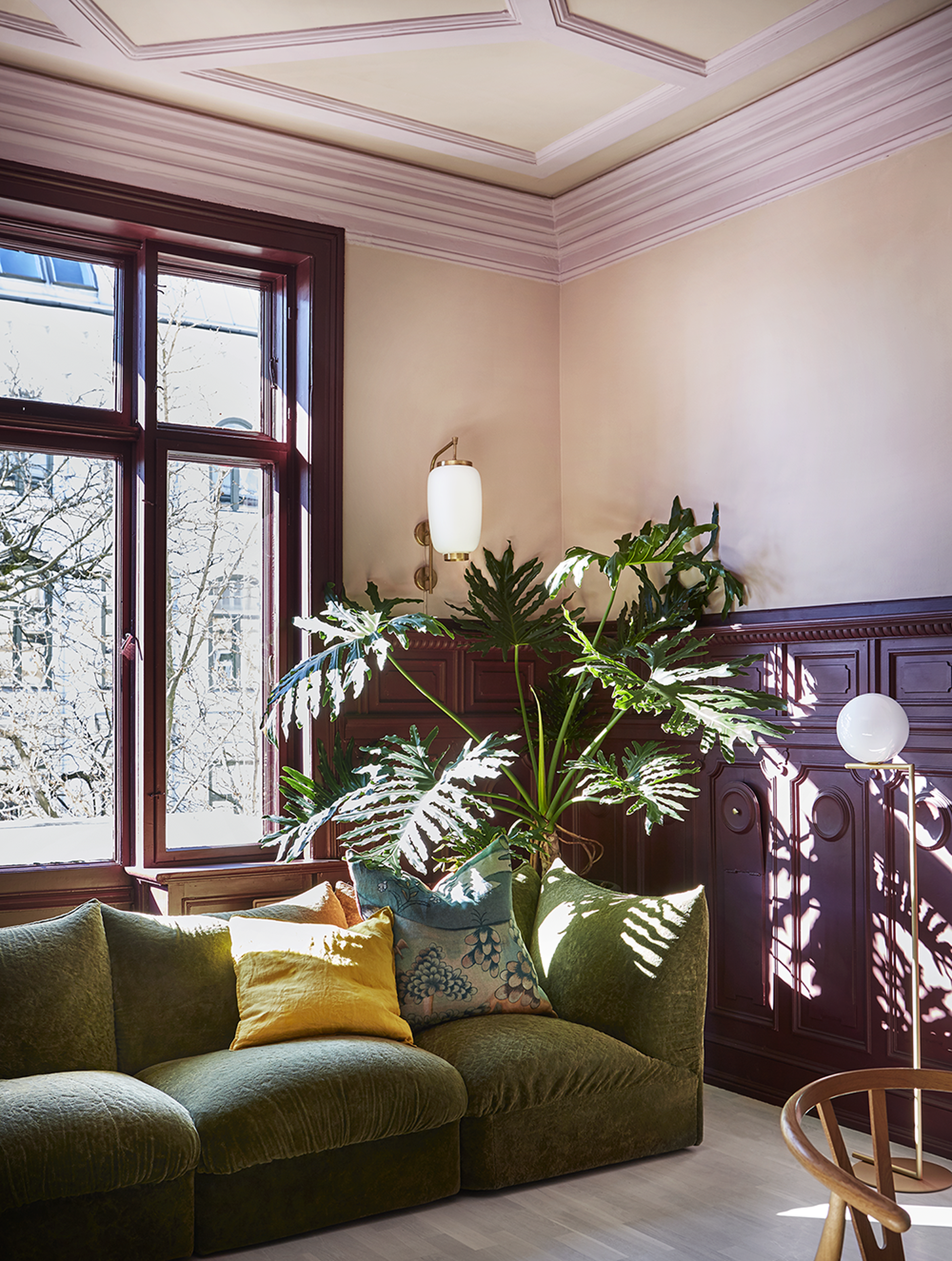 Stue med beige, rosa og lilla på vegger, tak og vinduskarm. Grønn sofa og stor grønnplante. Bilde.