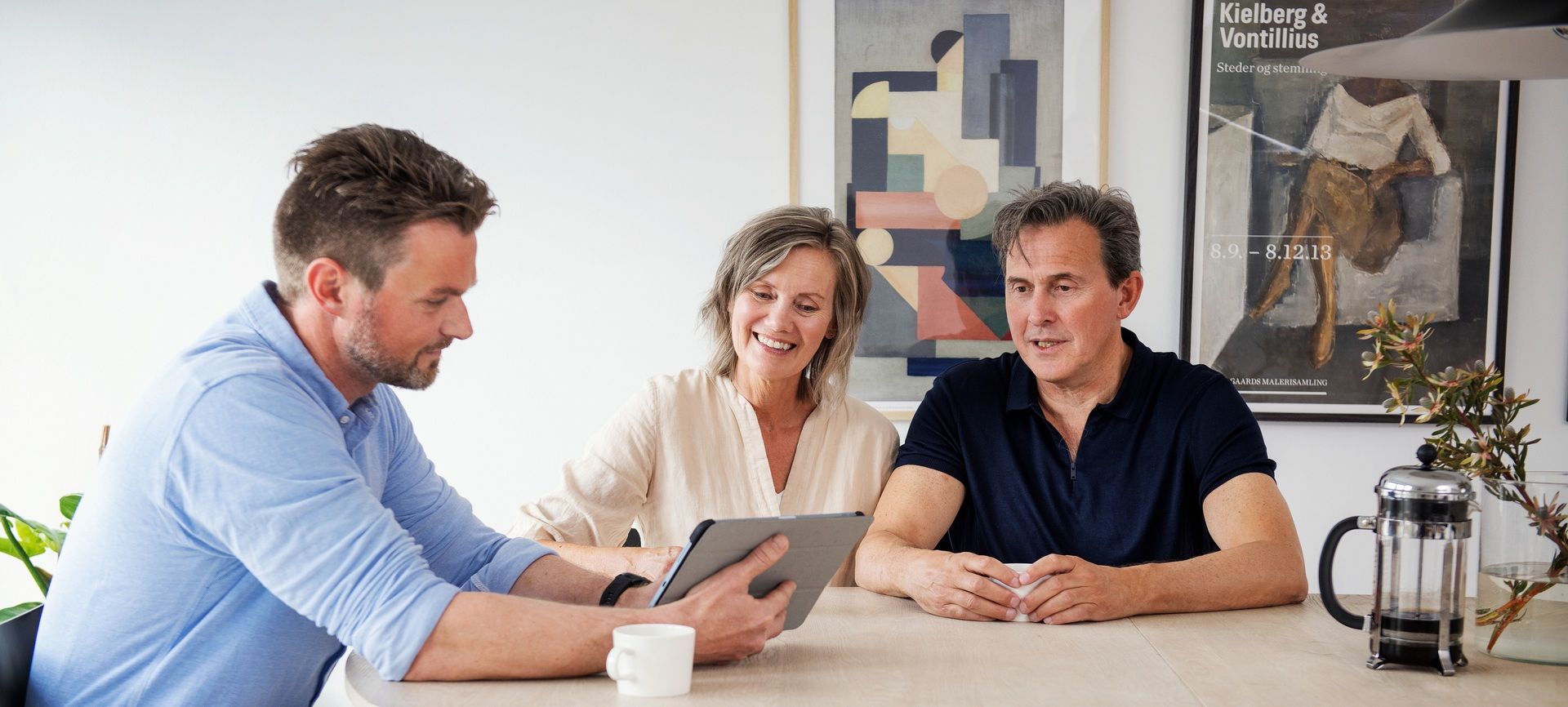 Par sitter sammen ved et bord i møte med en eiendomsmegler som viser frem noe på en tablet. Foto.