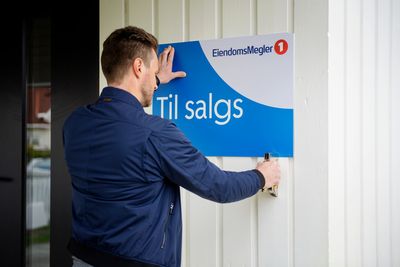 Foto av en mann som stifter fast et blått og hvitt til salgs-skilt fra EiendomsMegler 1 på en hvit husvegg.