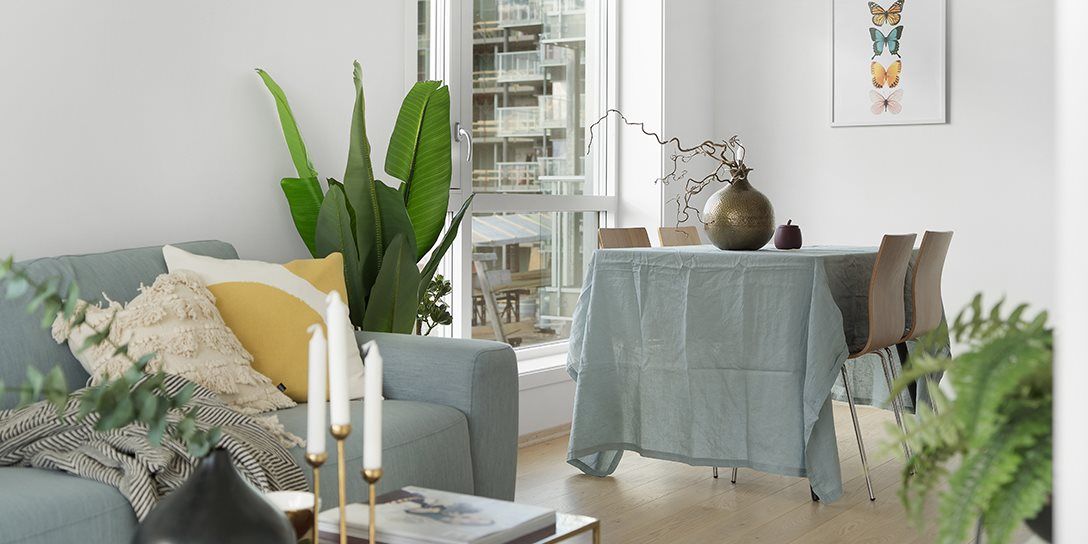 Stue med hvite vegger og lys blå sofa, spisebord og vindu med utsikt til naboblokk. Bilde.
