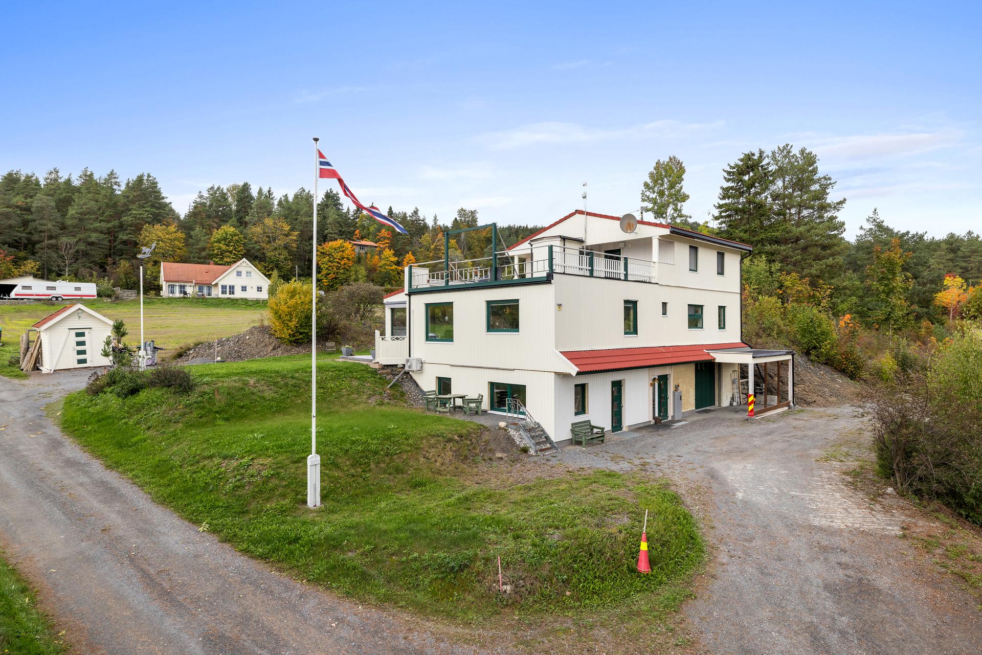 Frittliggende enebolig over tre plan med integrert garasje, separat boenhet i toppetasjen og landlig beliggenhet. Boligen sett fra adkomstveien, med inngang til underetasjen, gruset gårdsplass og takterrasse i andre etasje.. Bilde nummer 2 av 3