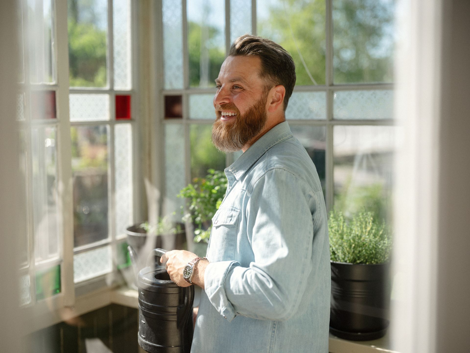 Foto av mann med skjegg som står i en vinterhage i et gammelt hus. I hånden holder han en vannkanne, og det er pyntet med grønne planter. 