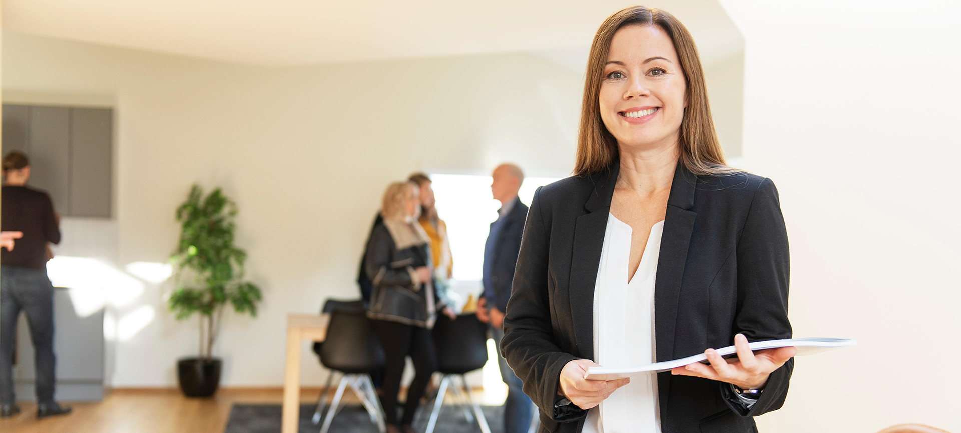 Kvinnelig megler på visning 1920x866.png Foto av en megler på visning som ser inn i kamera. I hånden har hun salgsoppgaver, og i bakgrunnen er det folk som kikker på boligen.