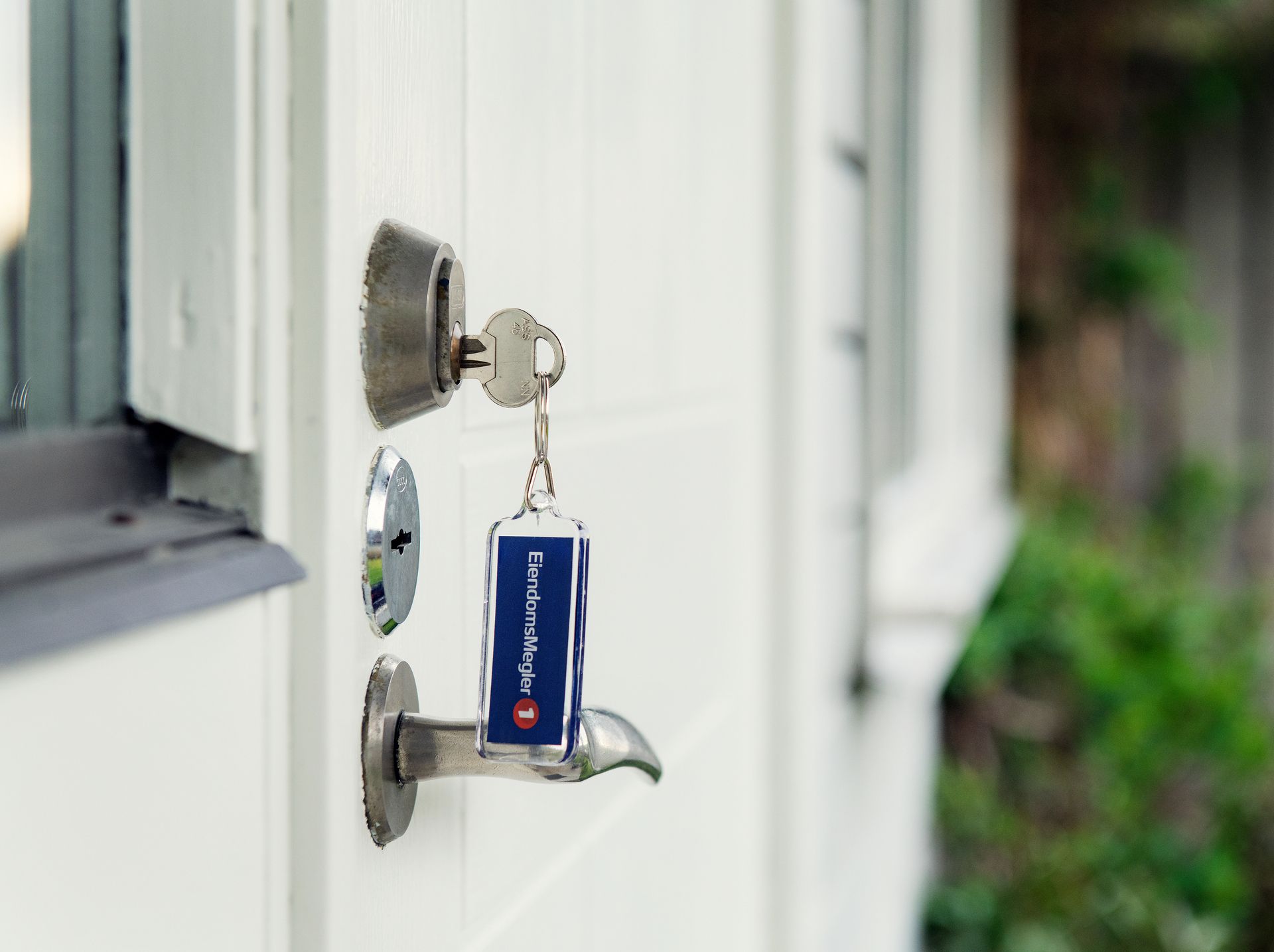 Nøkkel står i låsen på en hvit dør. Blå nøkkelring med EiendomsMegler 1-logo. Foto
