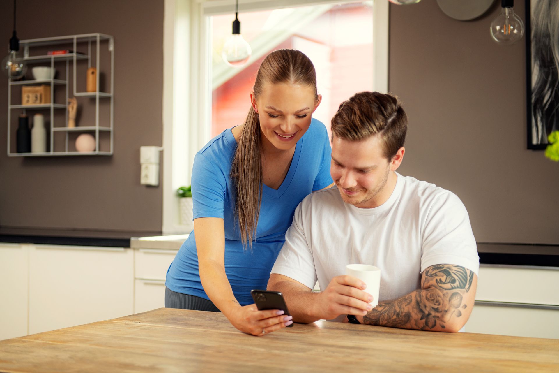 Par ved et bord, mann holder i en kaffekopp og dame viser frem noe på en mobil. Foto.
