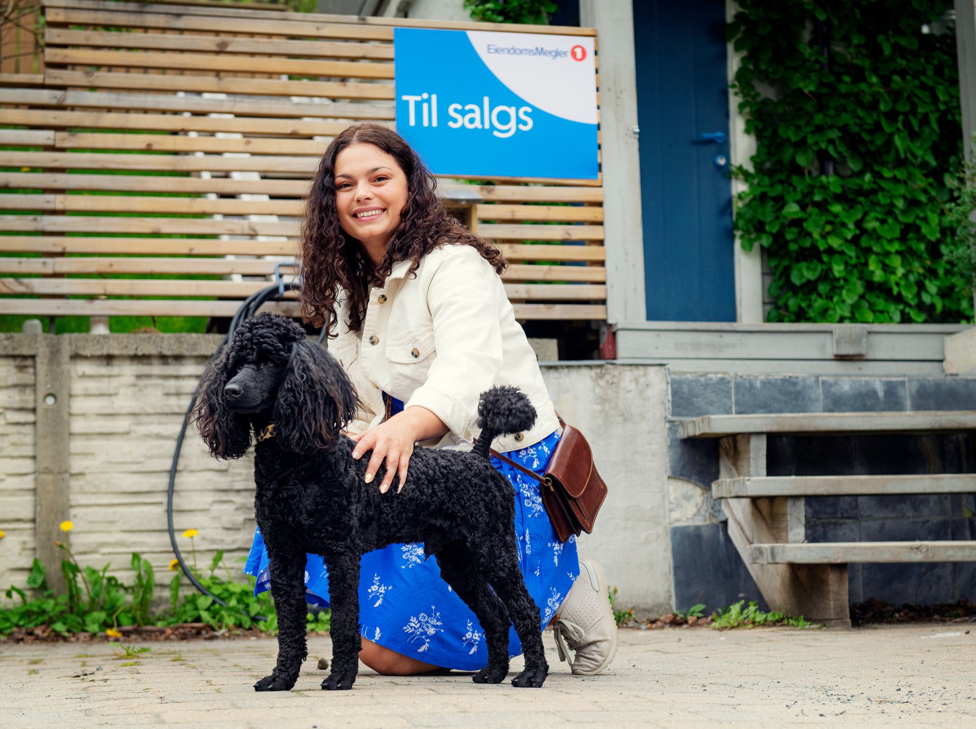 Kvinne sitter på huk sammen med en hund foran et hus med til salgs-skilt. Foto.
